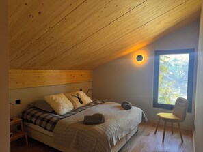 Ein Schlafzimmer mit schrägen Wänden, einem Bett mit karierter Decke, einem Stuhl und einem Fenster mit Blick auf Bäume.