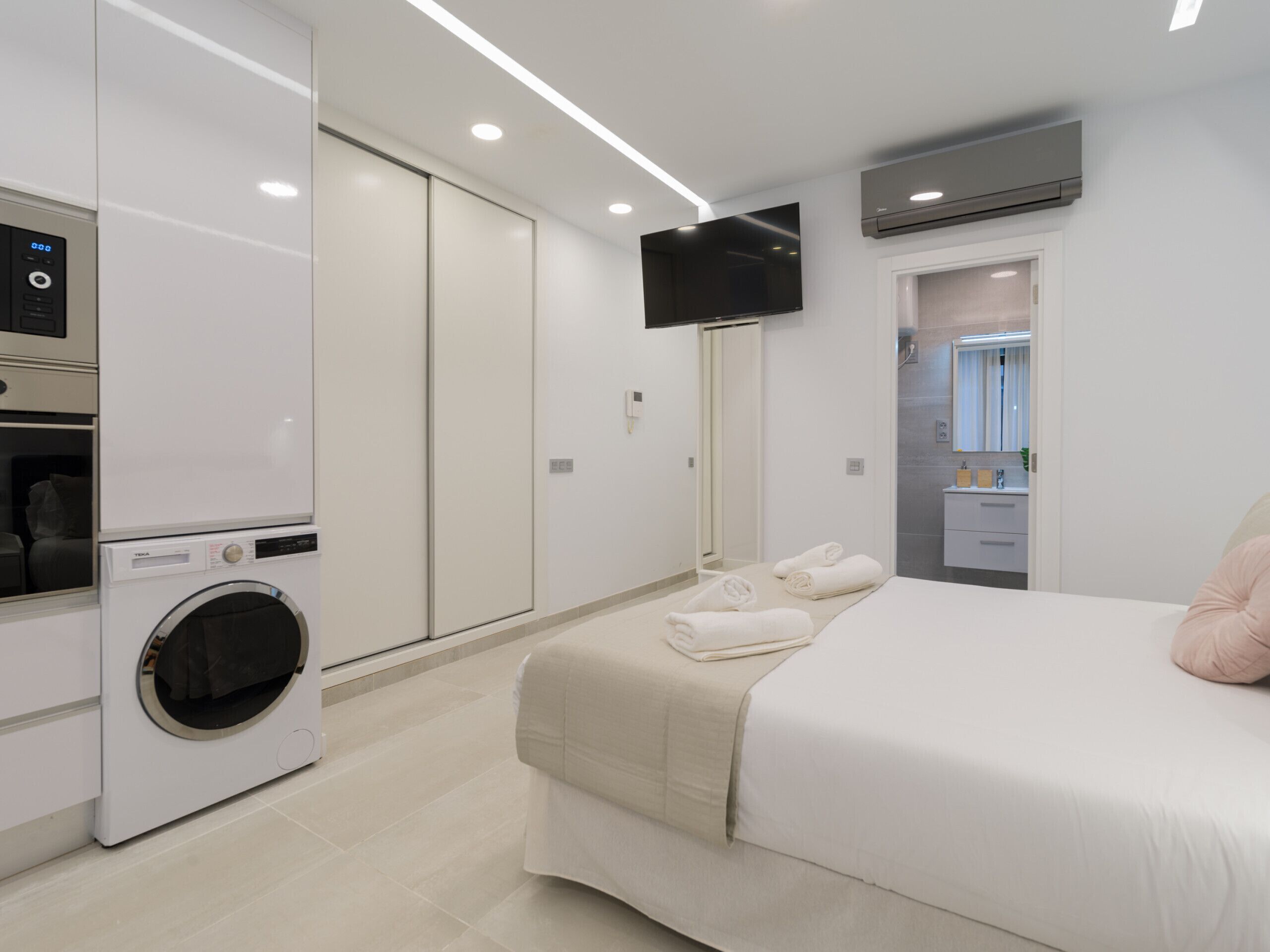 A modern hotel room with a bed, a washing machine, a TV, and a bathroom visible through an open door.