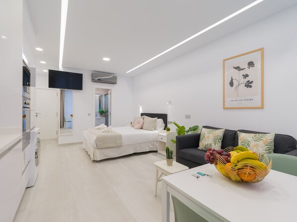 A modern hotel room with a bed, a sofa, a small table with fruit, a TV, and a framed picture on the wall.