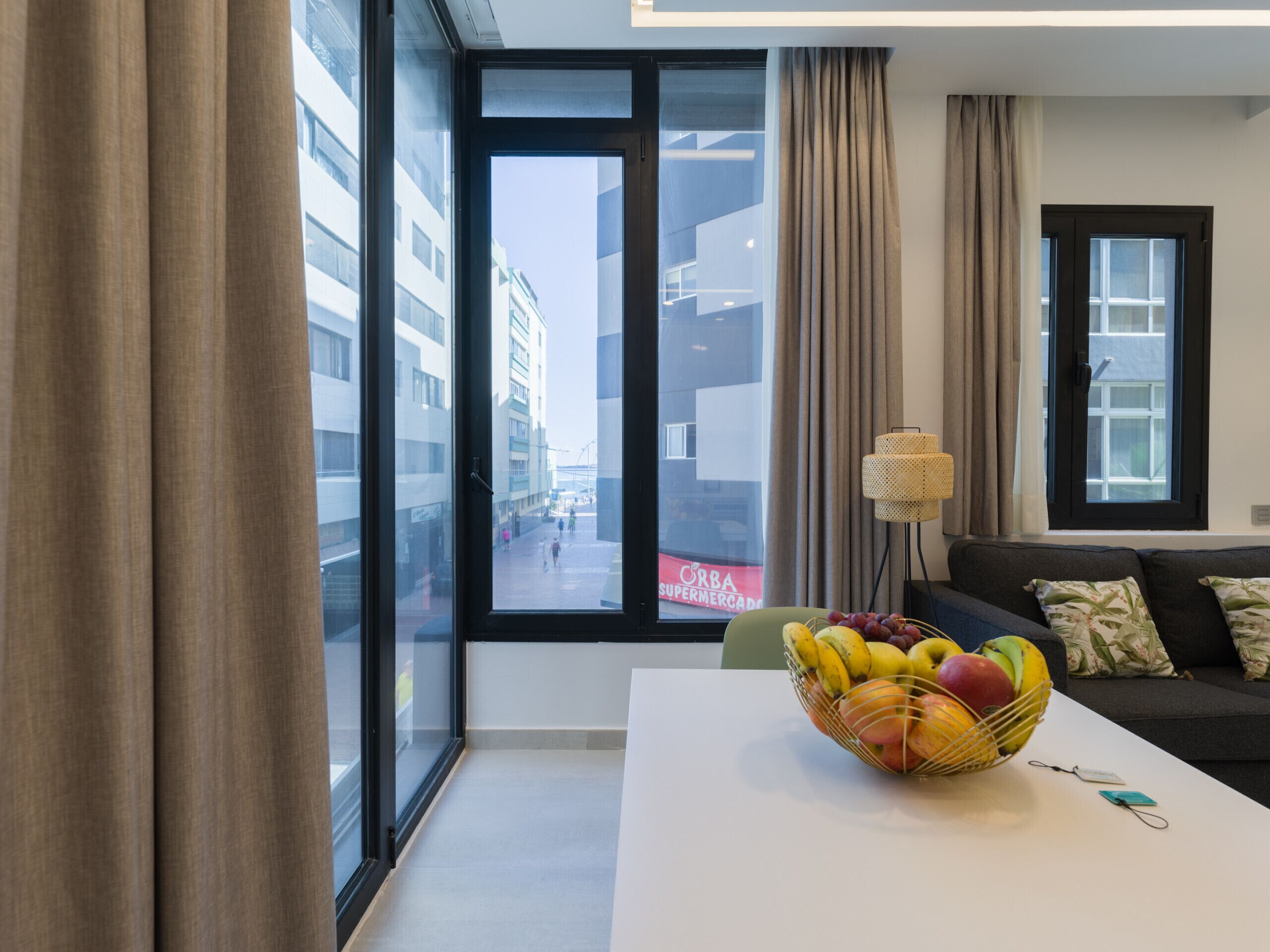 Ein modernes Wohnzimmer mit großem Fenster, einer Couch, einem Tisch mit Obstschale und einer Lampe.