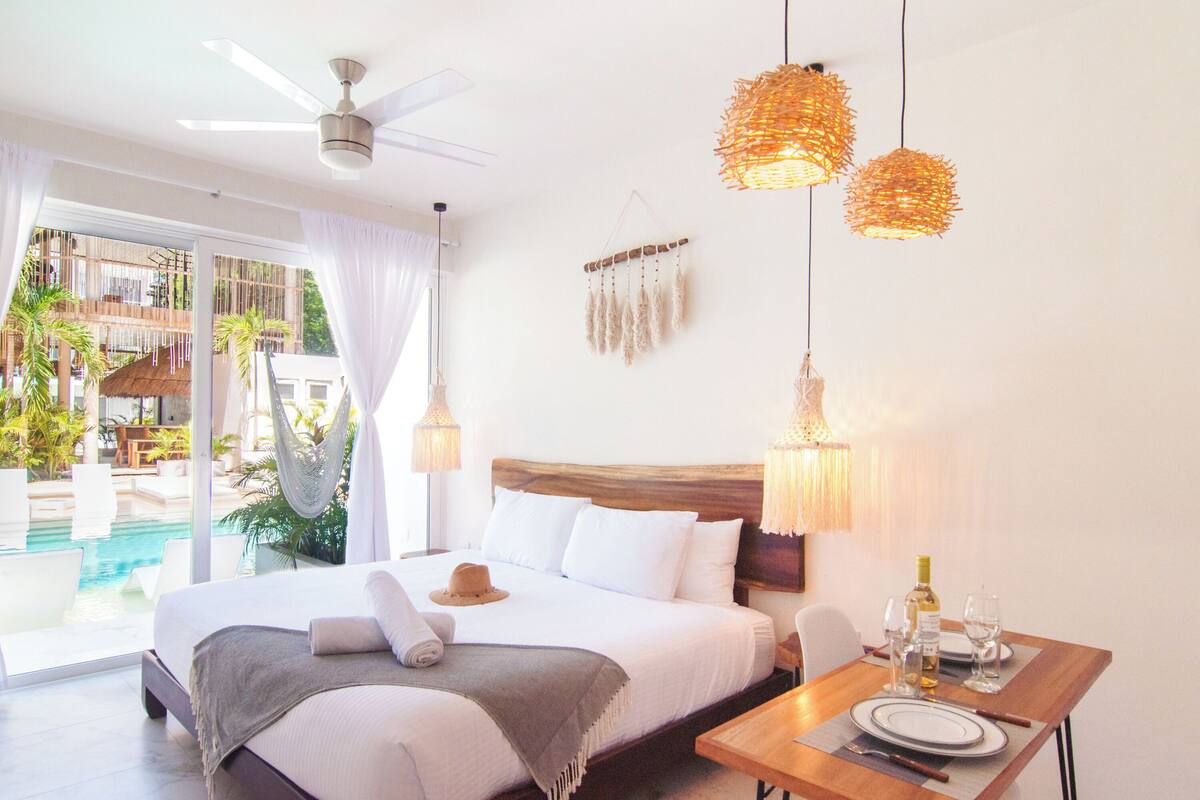 A bedroom with a bed, a dining table with plates and glasses, a ceiling fan, and a view of a pool and outdoor area.
