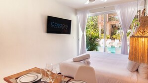 Una habitación de hotel con una cama, una mesa de comedor con platos y vasos, un televisor en la pared y vistas a una piscina y zonas verdes al exterior.