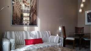 A living room with a patterned sofa, a coffee table, a dining table with chairs, and a painting of a building on the wall. - Vieux-Old Farnham Appart Condotel1 (Farnham)