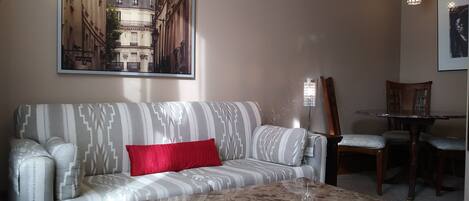 Una sala de estar con un sofá estampado, una mesa de centro, una mesa de comedor con sillas y un cuadro de un edificio en la pared.