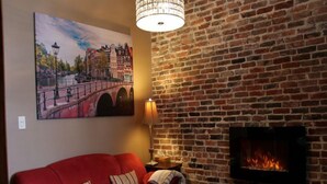 A cozy room with a red sofa, a fireplace, a brick wall, and a painting of a bridge. - Vieux-Old Farnham Appart Condotel1 (Farnham)