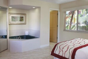 A hotel room with a bathtub, a bed with a red and white patterned bedspread, a framed picture on the wall, and a window with blinds.