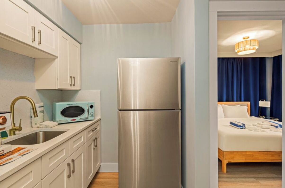 A compact hotel room with a kitchenette, a bed with a wooden headboard, and a blue curtain.