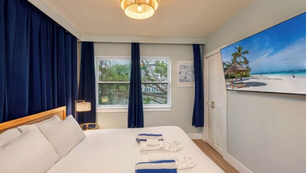 Una habitación de hotel con una cama, una mesita de noche, una lámpara, una ventana con cortinas y un cuadro que muestra una playa.