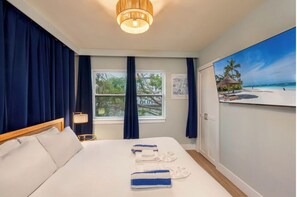 A hotel room with a bed, a nightstand, a lamp, a window with curtains, and a painting of a beach scene.
