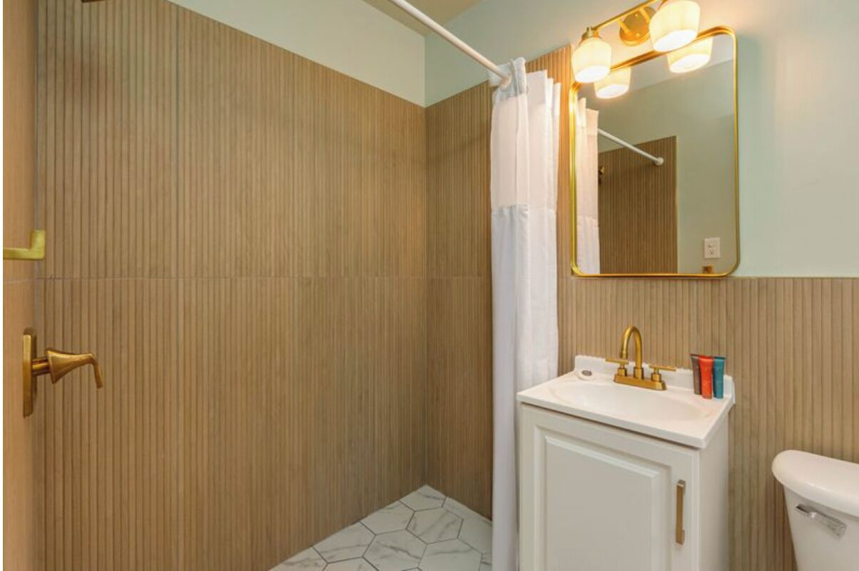 A bathroom with a wooden wall, a white sink with a gold faucet, a mirror with lights, and a shower curtain.