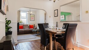 Ein Essbereich mit einem Holztisch, schwarzen Stühlen und einer Kristallleuchter. Im Hintergrund ist eine Couch mit roten Kissen zu sehen.