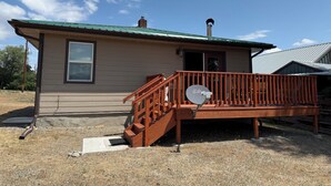 A small single-story building with a green metal roof, a wooden deck with a satellite dish, and a window.