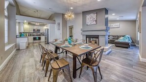A spacious living area with a dining table set for six, a fireplace, and a kitchen in the background.