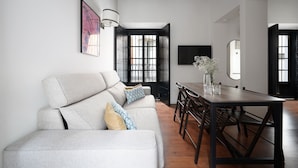 A modern living room with a sofa, dining table, and chairs. There is a large window with black frames, a wall-mounted TV, and a decorative vase on the table. - Numa Seville Alegre (Seville)