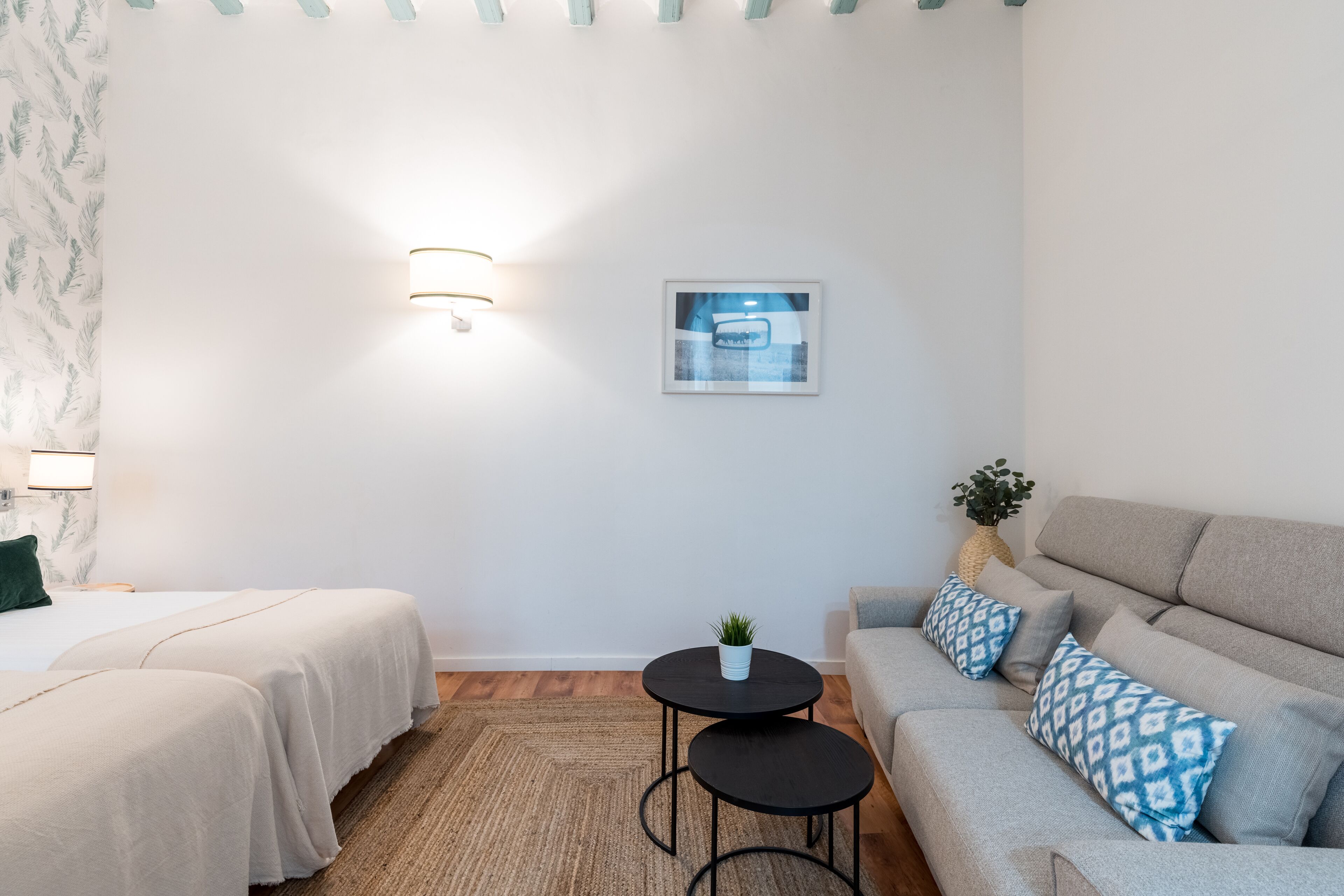 Ett modernt hotellrum med två sängar, en soffa, ett litet bord med en växt och en inramad tavla på väggen.