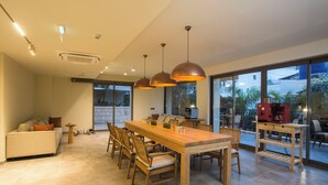 Un vaste espace dédié à la salle à manger, avec une longue table en bois et des chaises, des luminaires suspendus et une machine à café posée sur un présentoir en bois.