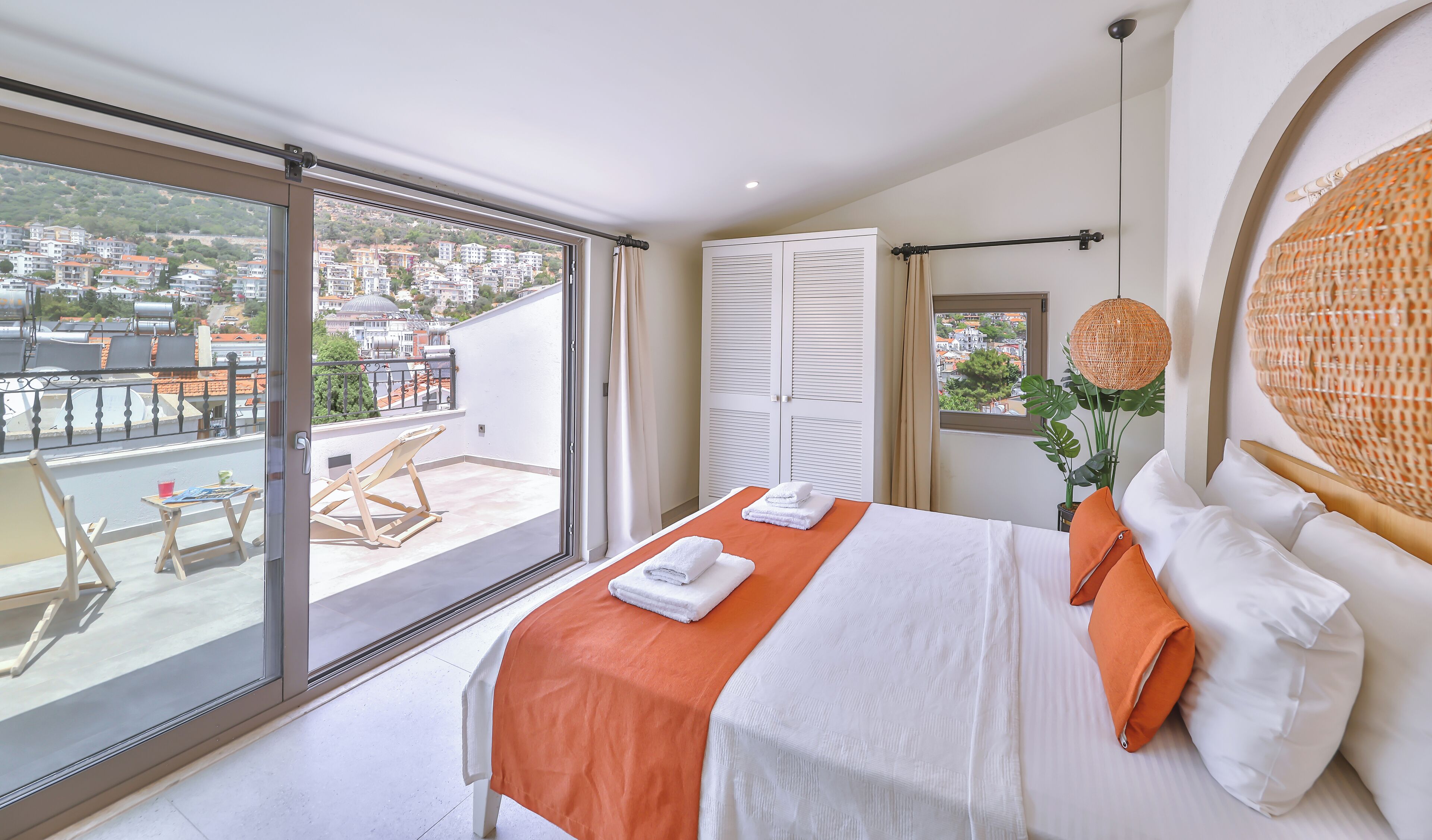 Habitación de hotel con balcón, cama con ropa de cama blanca y naranja, un ventanal grande y vistas a la ciudad.