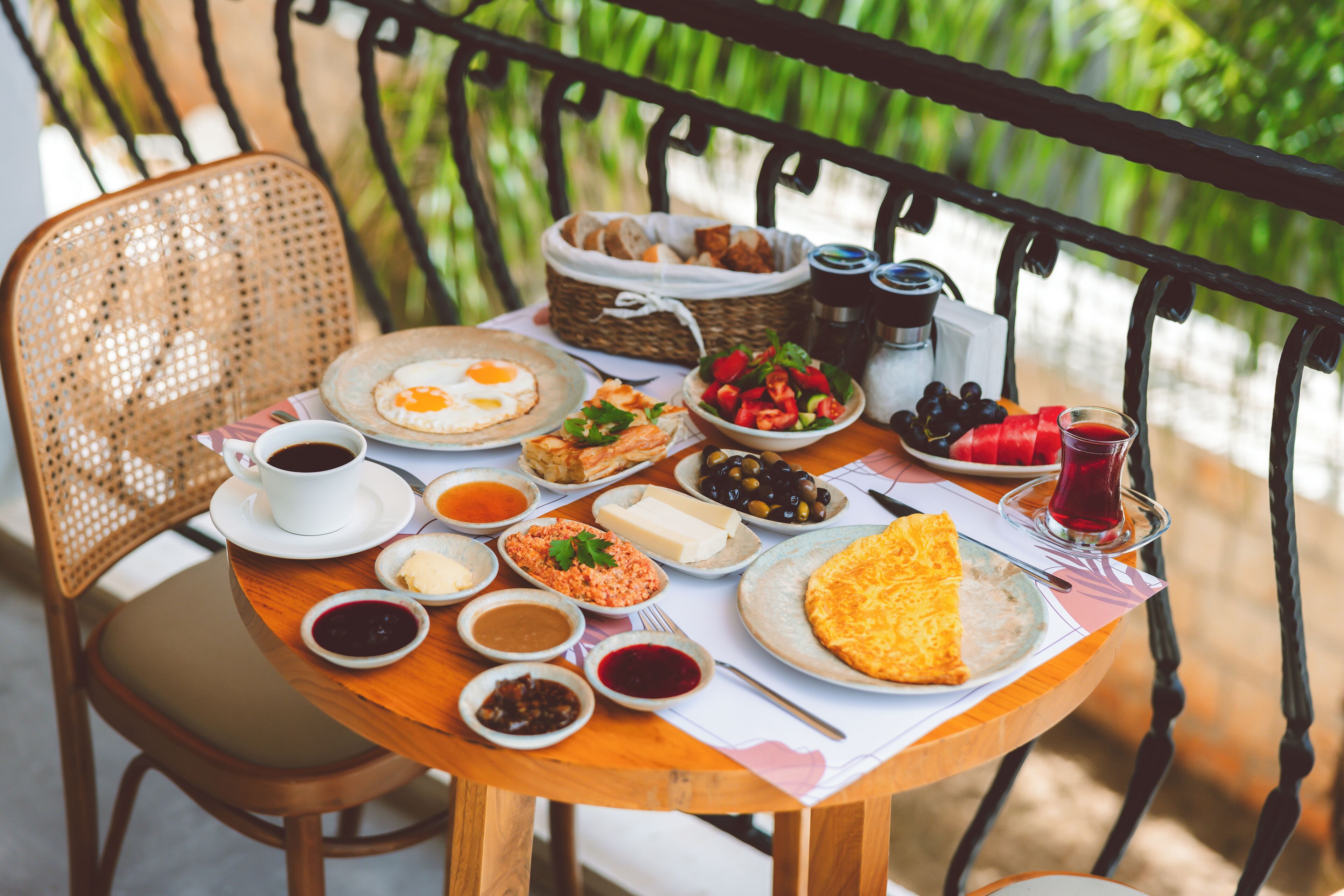 A round wooden table with a variety of breakfast items including eggs, bread, and fruits, set on a balcony with a metal railing.