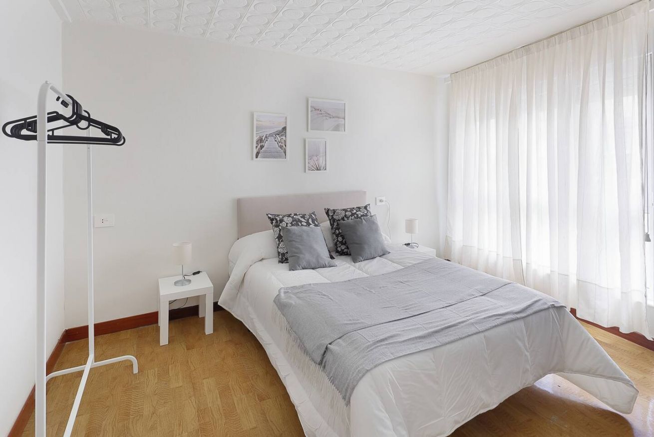 Un dormitorio con cama, mesita de noche, lámpara, perchero y ventana con cortinas.