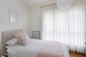 A bedroom with a bed, a headboard, a radiator, and a window with sheer curtains.