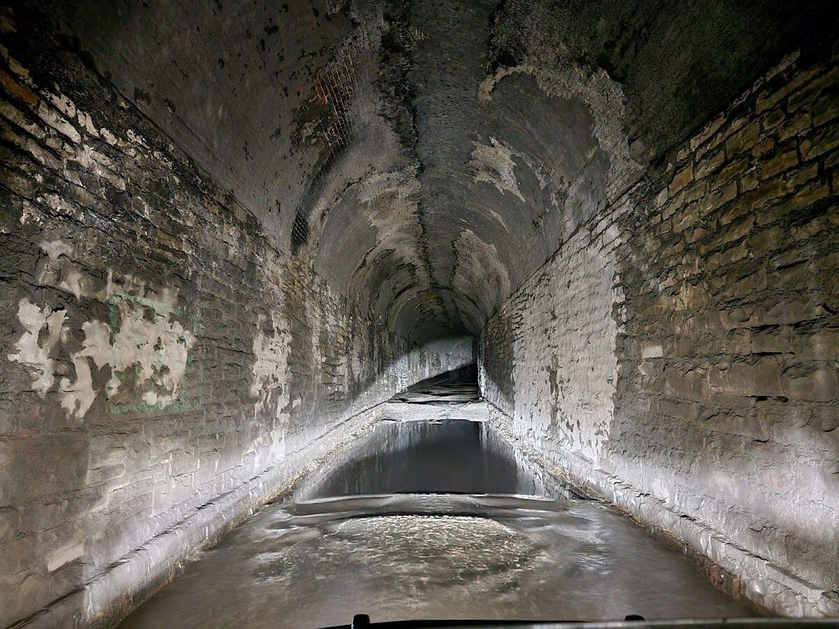 Un tunnel aux murs de briques et au sol en béton, qui mène à une zone plus lumineuse à l’extrémité.