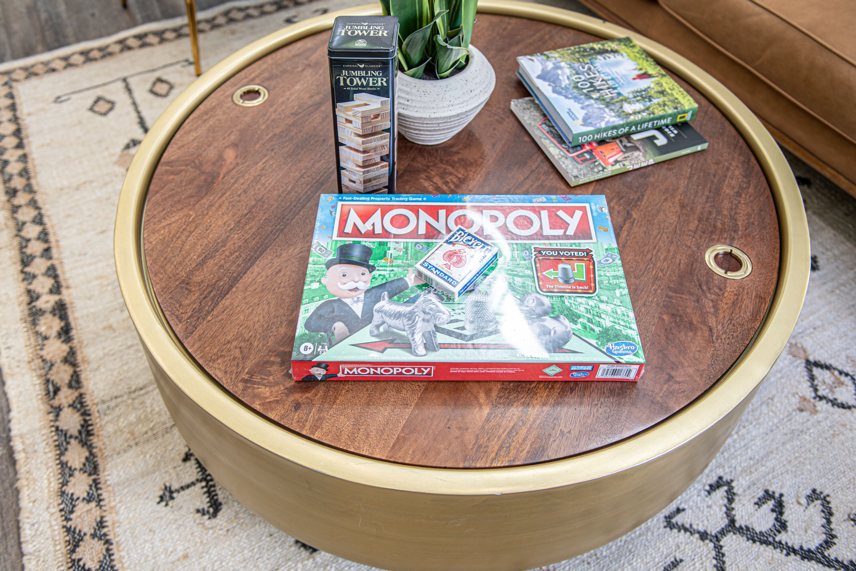 Een ronde houten salontafel met een Monopoly spelbord, een JUMBLING TOWER doos en een LIFEHOKES boek erop.