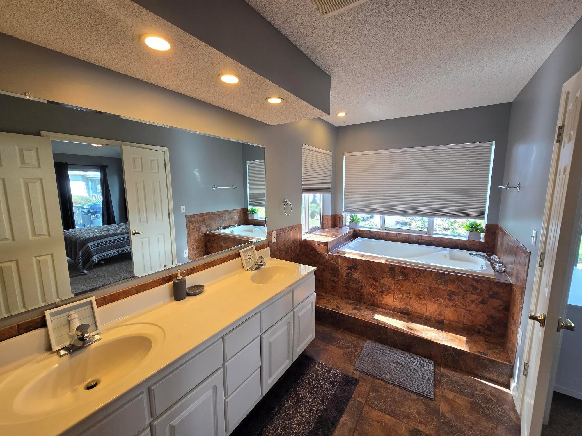A modern bathroom with a double sink, a large mirror, a bathtub, and a window with blinds.