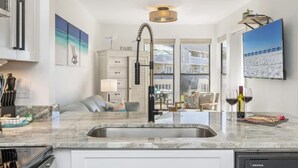 Una cocina moderna con encimera de mármol, pileta de acero inoxidable y lavavajillas integrado. Hay una zona de comedor con mesa y sillas, y un ventanal que ofrece vistas al exterior.