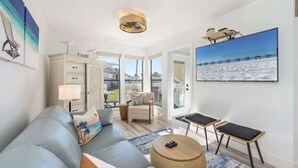 Una sala moderna con un sofá, una mesita de centro y un televisor. Se ve la playa a través de amplias ventanas.