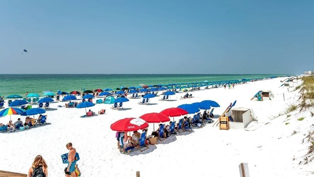 Una playa con hileras de sombrillas azules y rojas, arena blanca y el océano al fondo.