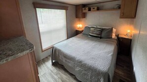 A compact bedroom with a bed, bedside tables, and a window with blinds. - Campground Tiny Home #46 (Huntsville)