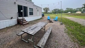 A rustic outdoor patio with a wooden table and benches, a fire pit, and blue chairs. - Campground Tiny Home #46 (Huntsville)
