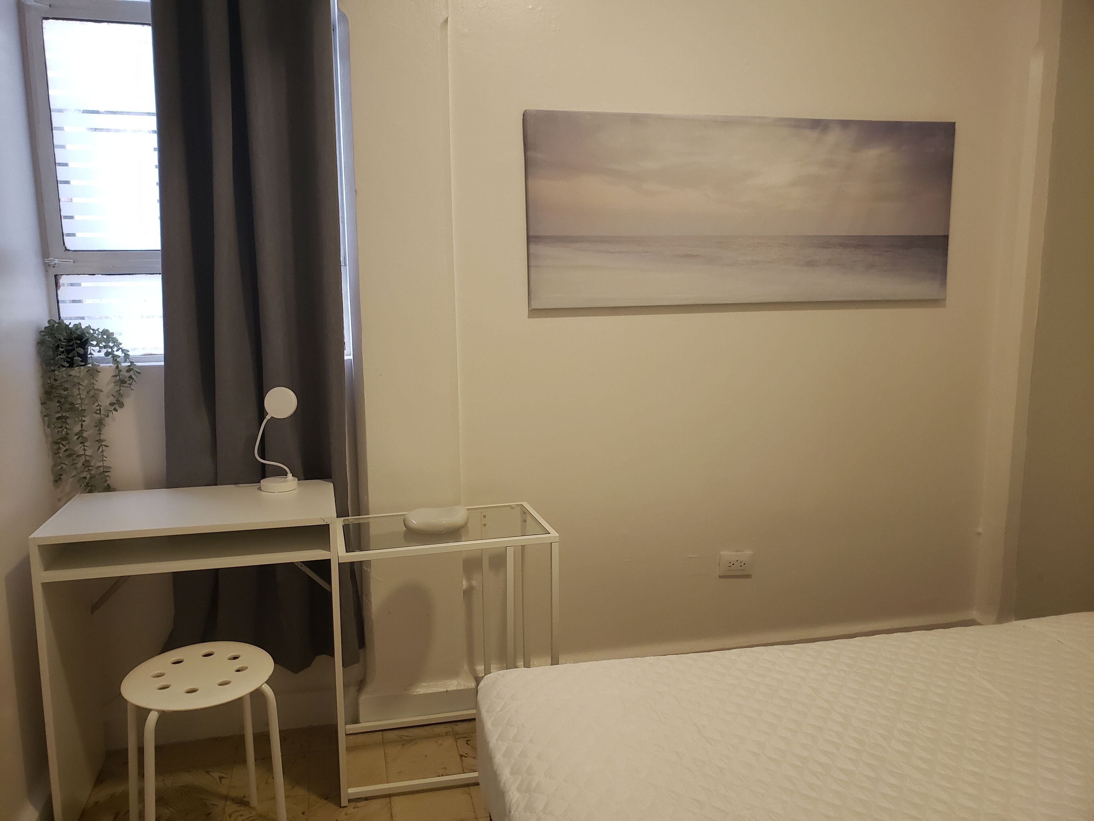 A bedroom with a bed, a desk with a chair, a window with curtains, a wall-mounted painting, and a potted plant.