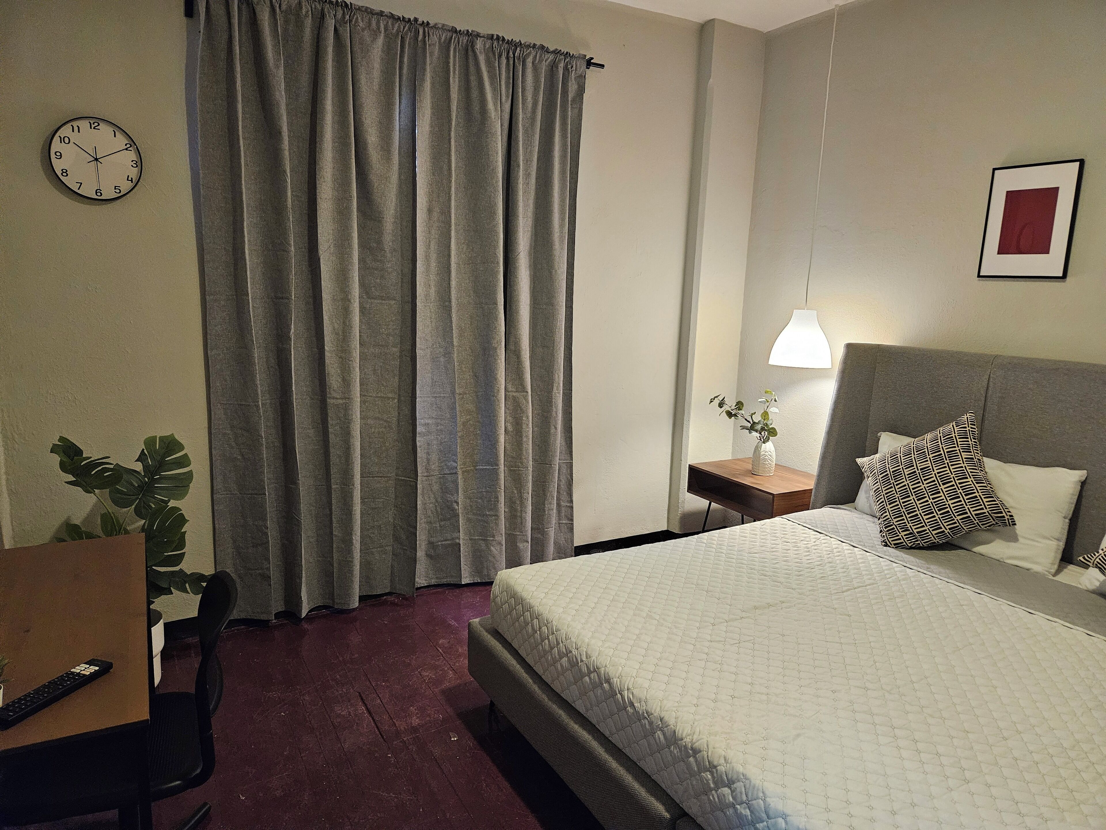 A hotel room with a bed, a desk with a chair, a plant, a clock, and a curtain.
