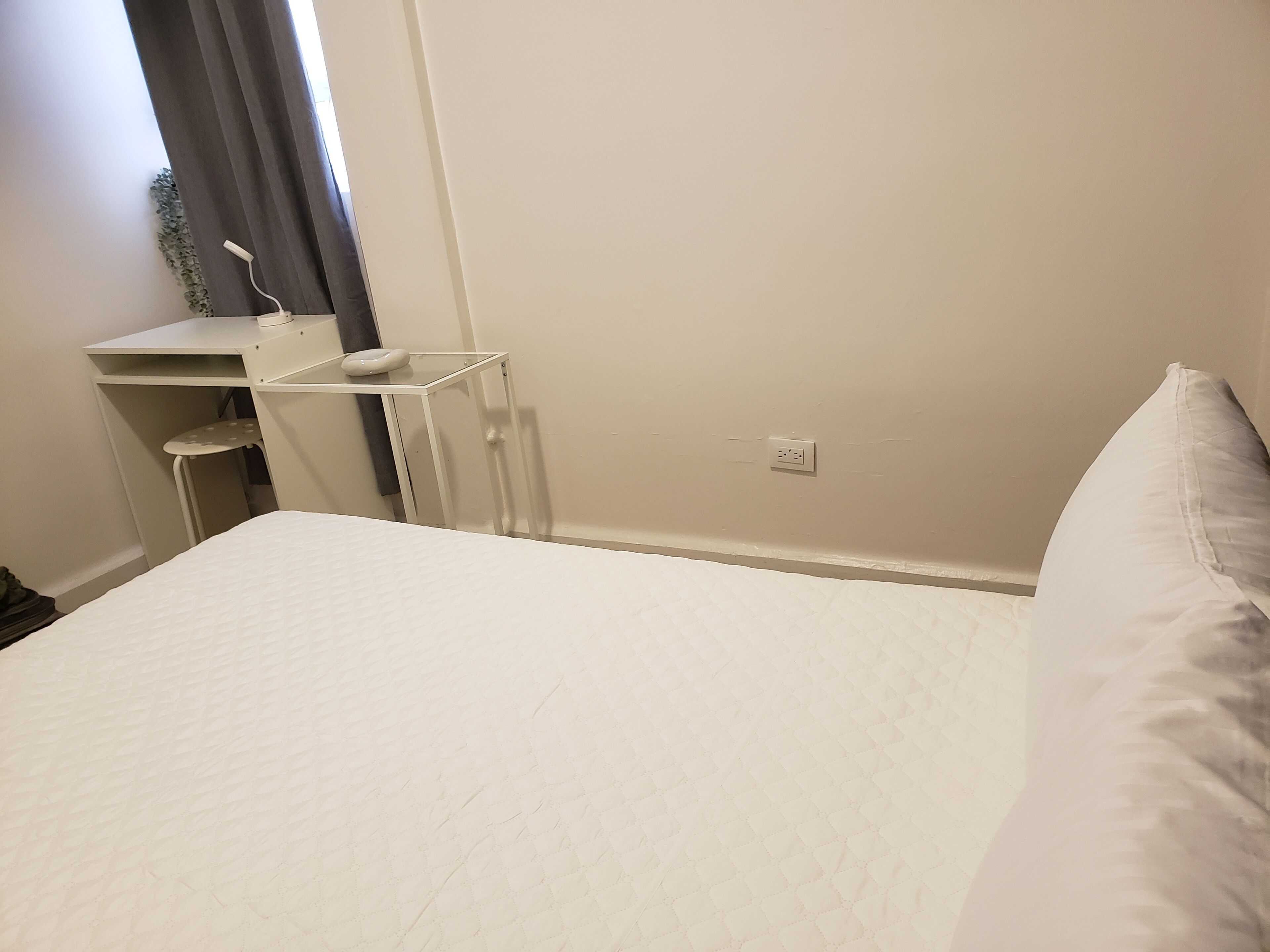 A bedroom with a bed, a desk, and a chair. There is a window with curtains, a wall socket, and a small plant on the desk.