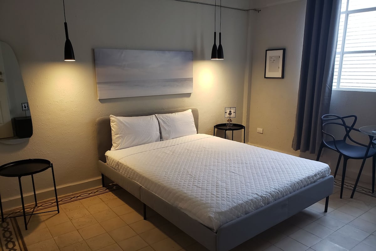 A neatly made bed with a quilted white duvet, a small bedside table with a lamp, a chair, and a framed picture on the wall.