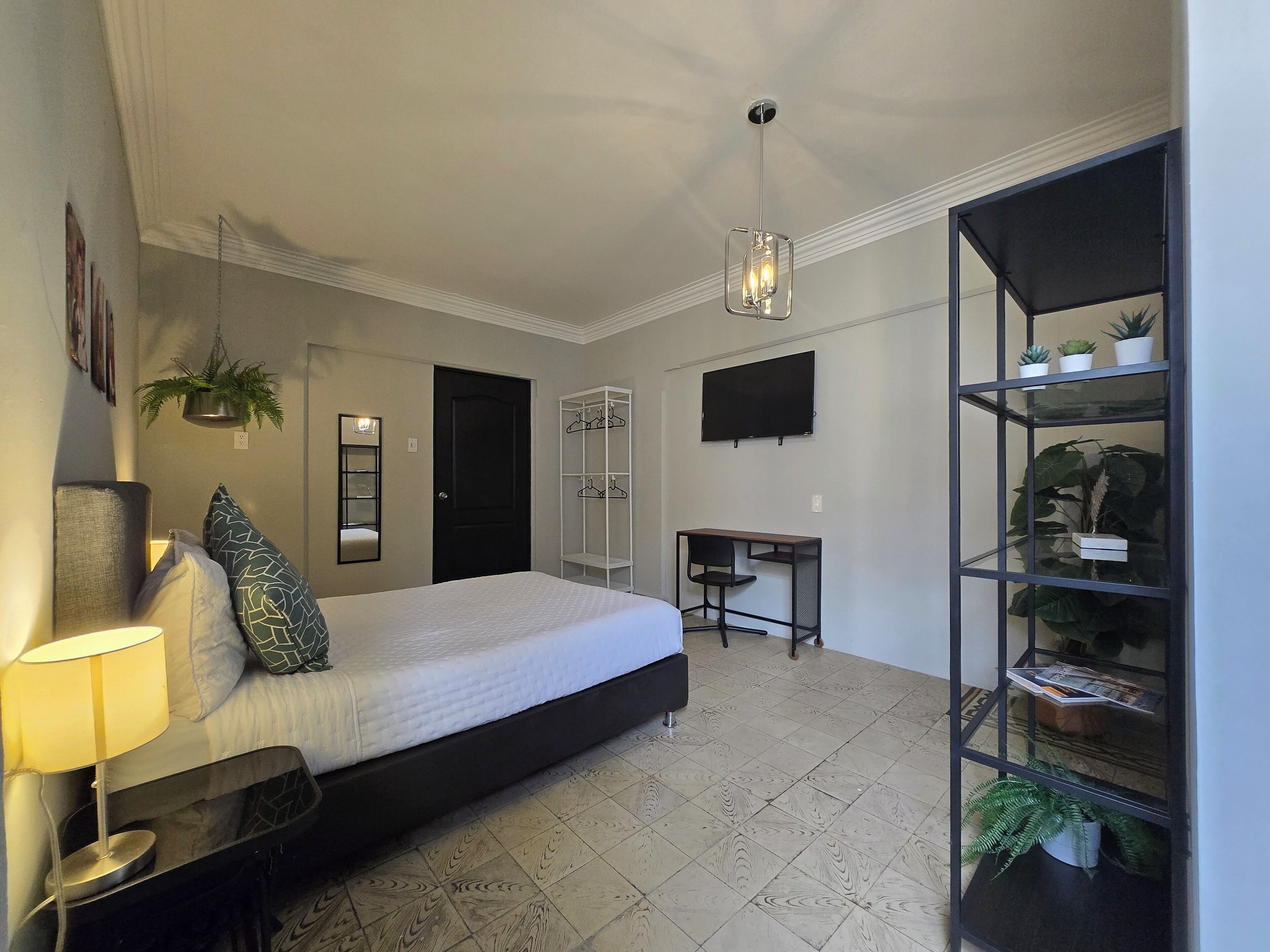 A bedroom with a bed, bedside table, lamp, hanging plant, wardrobe, desk, chair, and a TV on the wall.