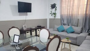 A modern living room with a glass dining table, wooden chairs, a grey sofa with blue cushions, a mounted TV, and a decorative plant.