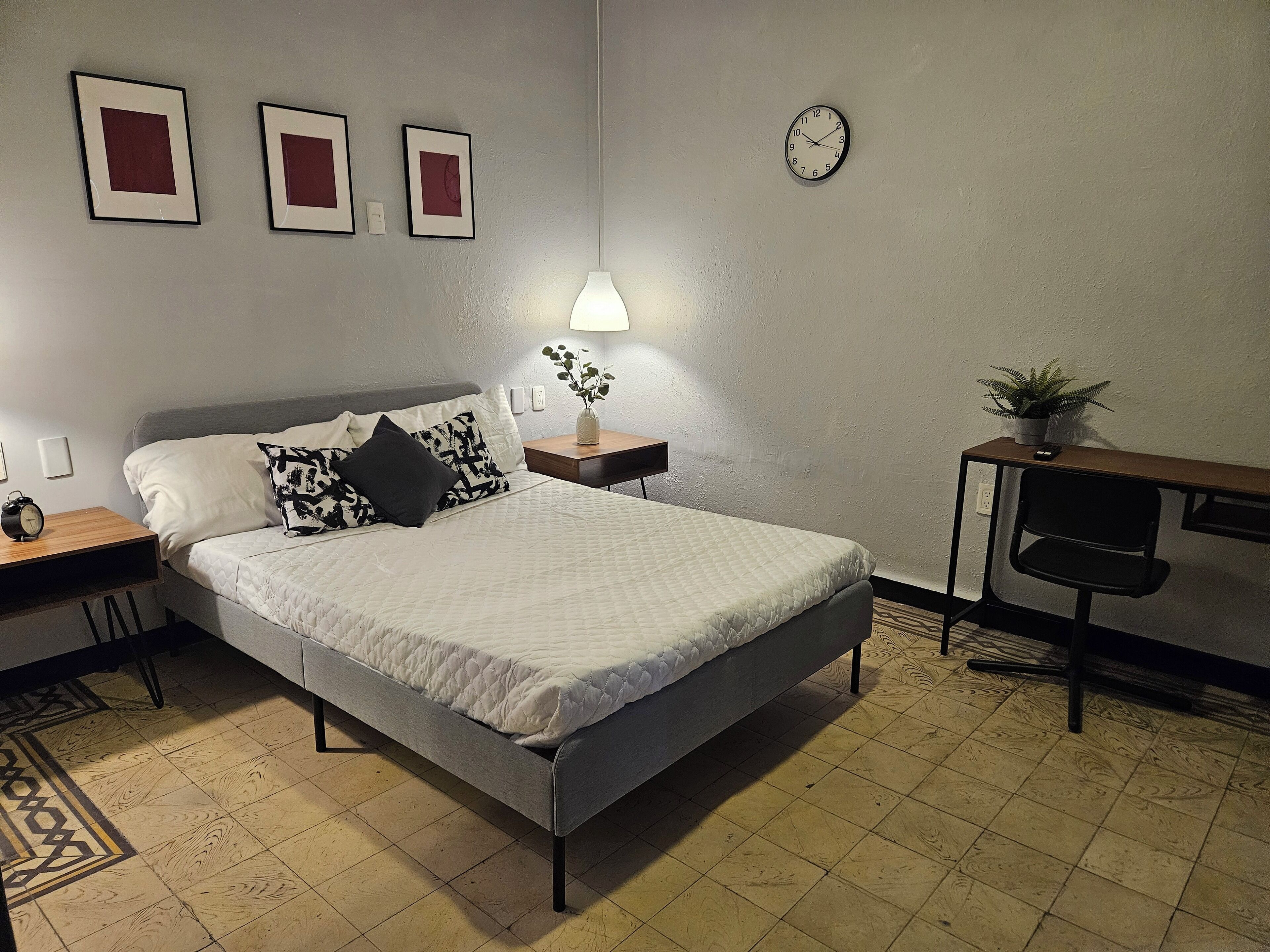 A bedroom with a bed, bedside tables, a desk, a chair, a clock, and framed pictures on the wall.