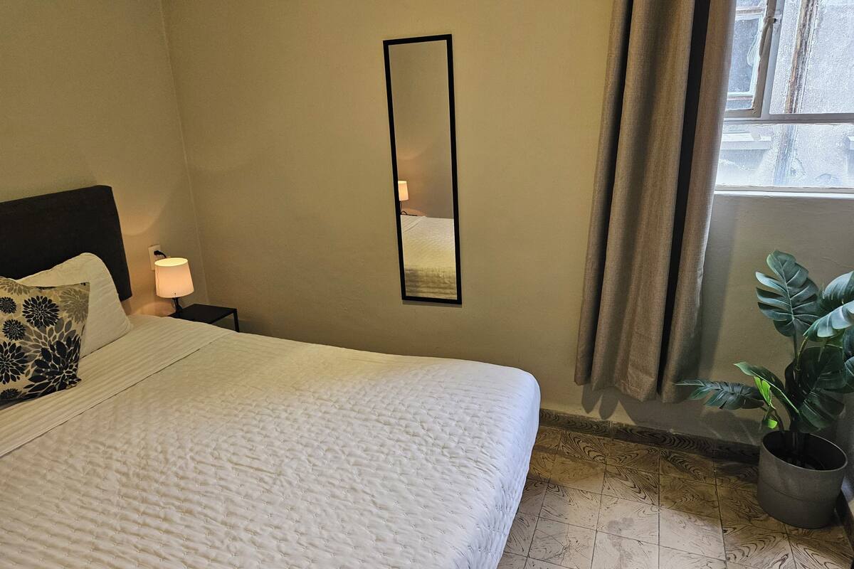 A neatly made bed with a white duvet, a bedside lamp, a mirror on the wall, and a potted plant by the window.