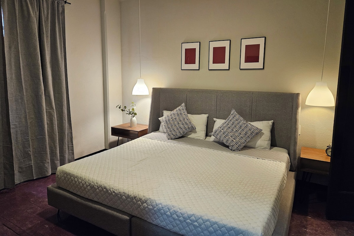 A neatly made bed with a quilted cover, a bedside table with a lamp, and a wall with three framed pictures.