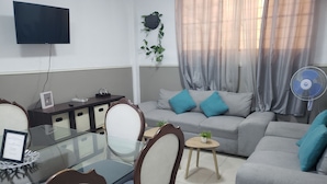 A living room with a glass-top dining table, a sofa with blue cushions, a grey couch, a TV mounted on the wall, and a standing fan.