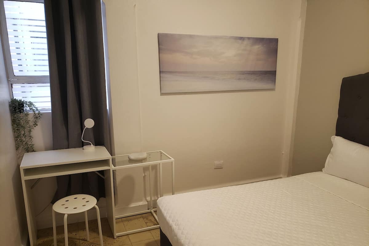A bedroom with a bed, a desk with a chair, a small stool, a radiator, and a wall-mounted painting.