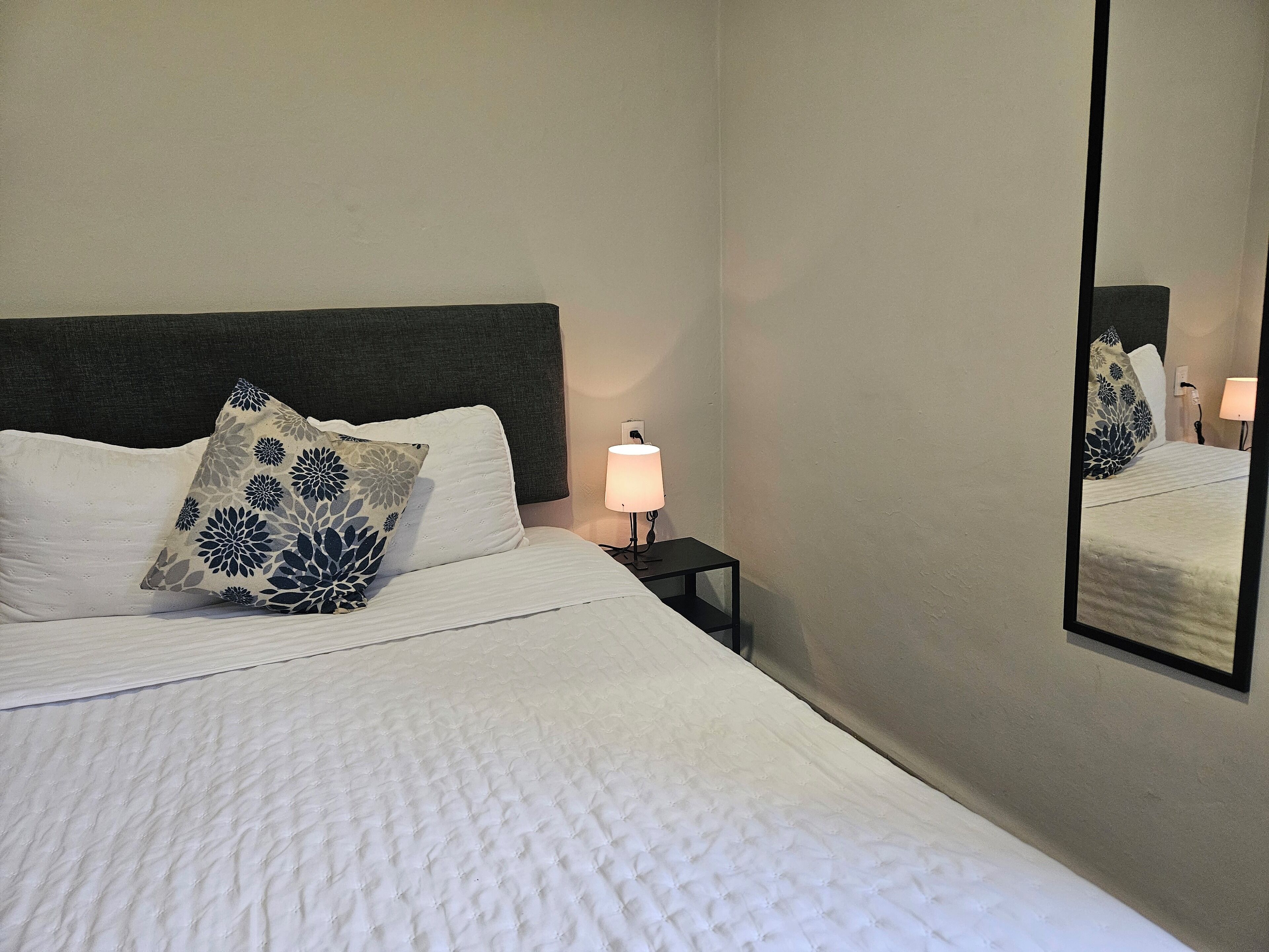 Una cama bien tendida con edredón blanco y almohada decorativa, una lámpara de noche y un espejo en la pared.