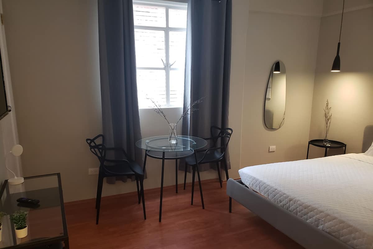 A bedroom with a bed, a round glass table with chairs, a small table with a vase, and a mirror on the wall.