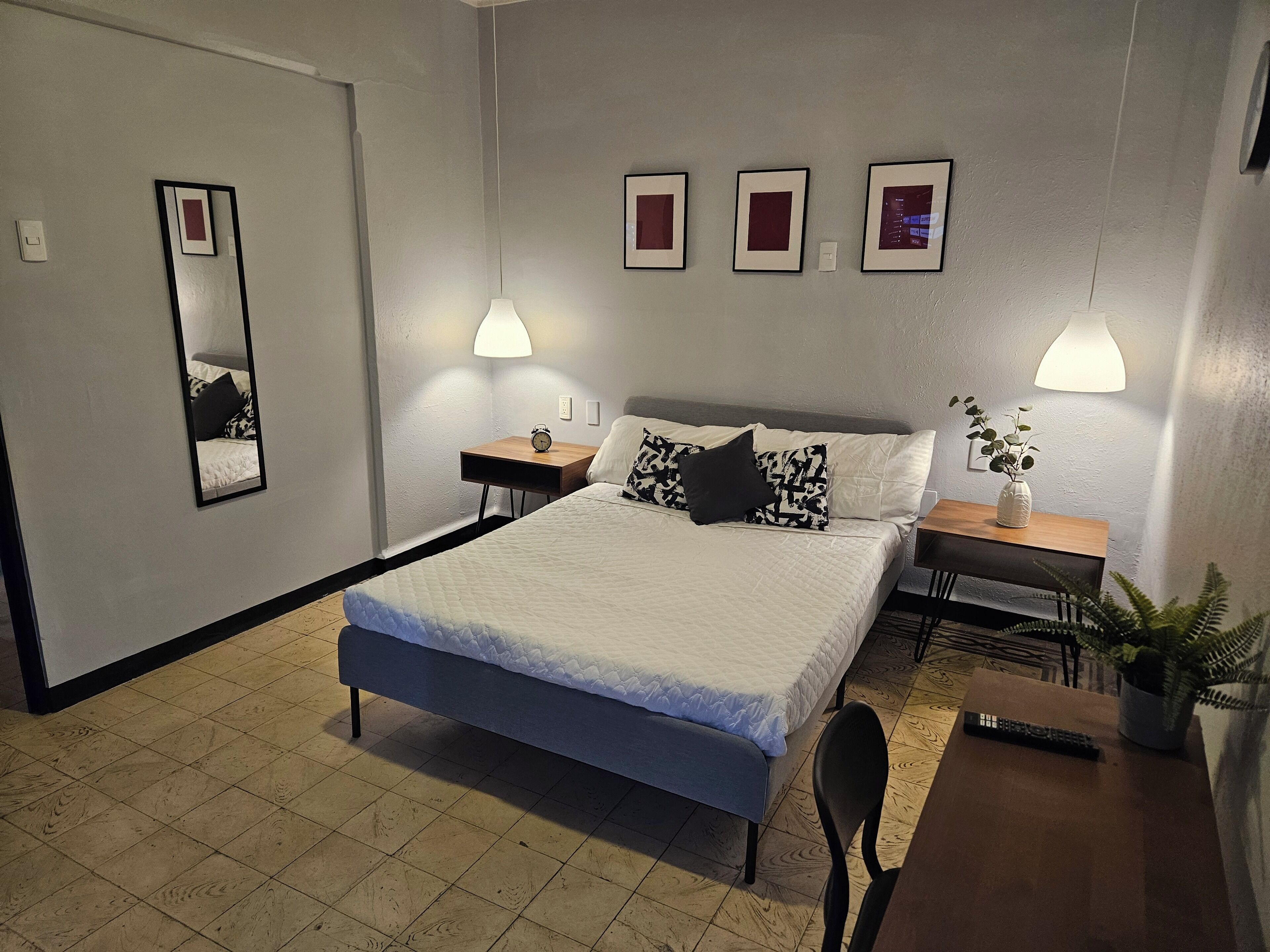 Habitació d'hotel ben ordenada amb llit, taules de nit, mirall i làmpades penjades a la paret.