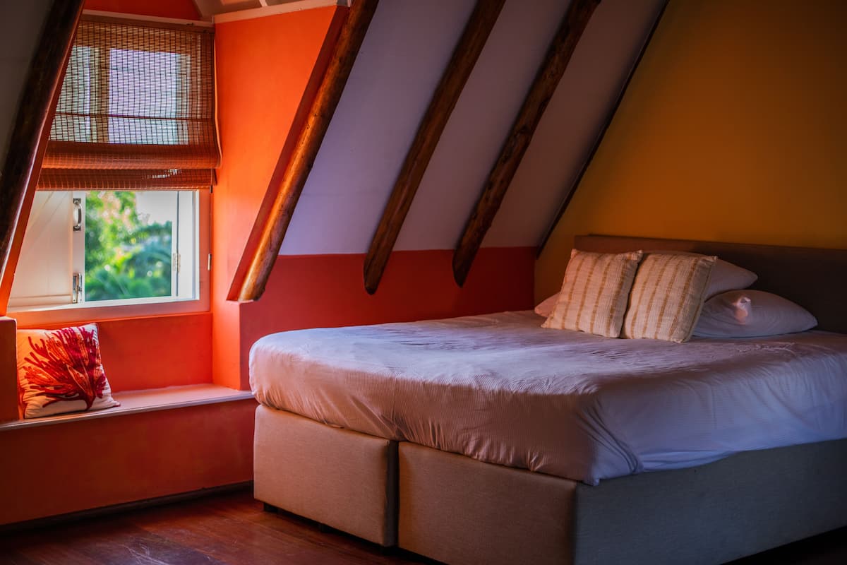 Een slaapkamer met een schuine kap, een bed met wit beddengoed, een raam met uitzicht op groen en een rode wand.