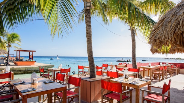 Een terras met houten tafels en rode stoelen, met uitzicht op een strand met palmbomen en de oceaan.