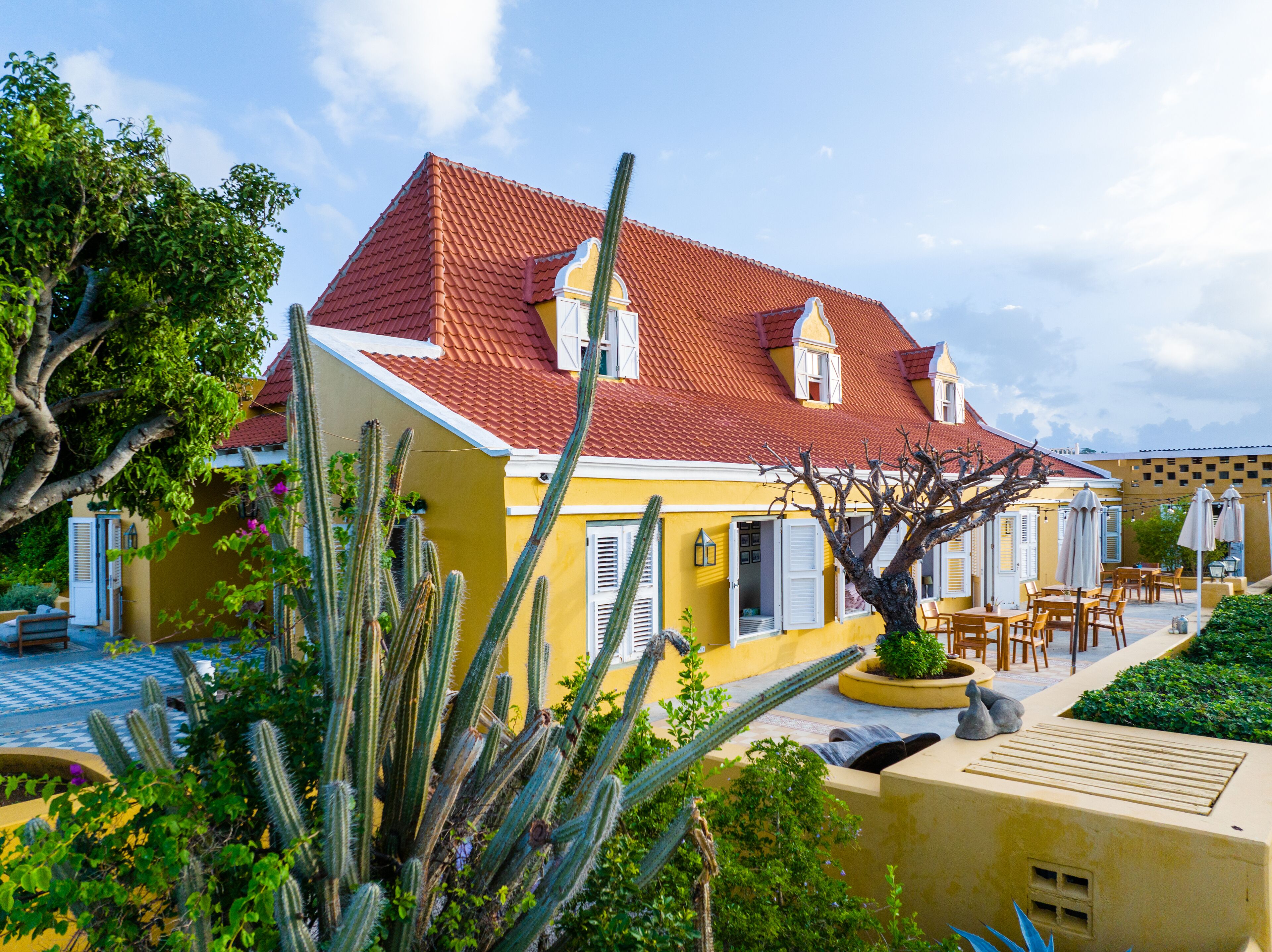 A yellow building with a red-tiled roof, white shutters, and a cactus garden.
