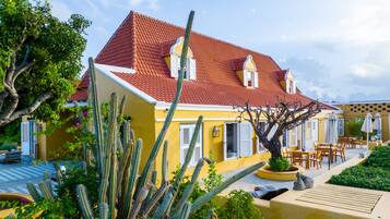 A yellow building with a red-tiled roof, white shutters, and a cactus garden.